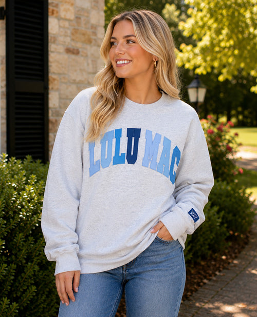A woman wearing a light heather gray sweatshirt with 'LULU MAC' printed across the chest in blue and navy, paired with blue jeans, standing outdoors in front of a stone building and greenery.