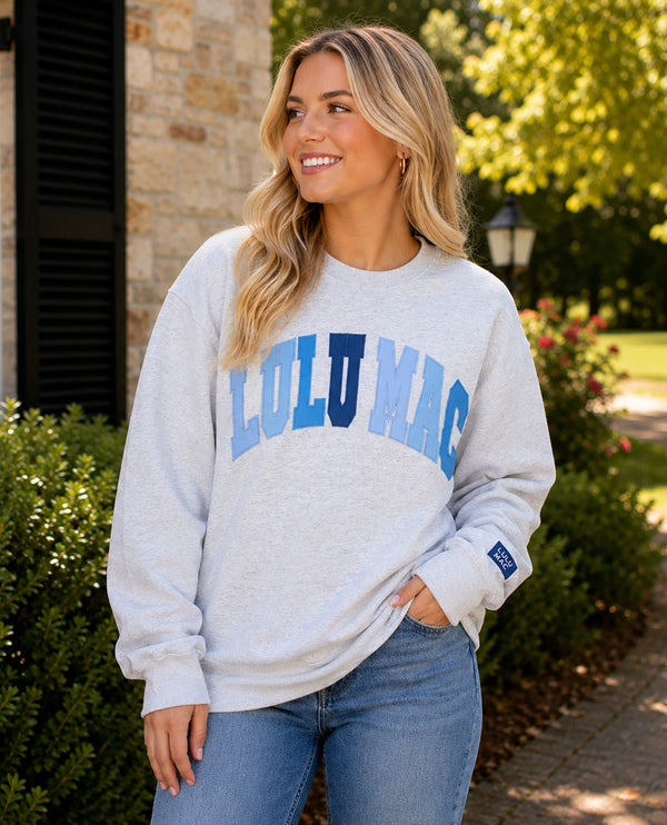 A woman wearing a light heather gray sweatshirt with 'LULU MAC' printed across the chest in blue and navy, paired with blue jeans, standing outdoors in front of a stone building and greenery.