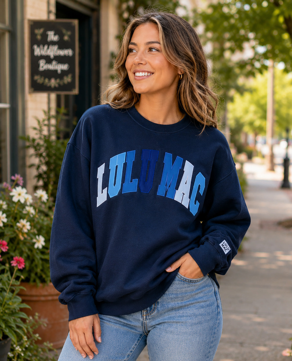 A woman wearing a navy blue crewneck sweatshirt with 'LULU MAC' printed in blue and light blue block letters across the chest, paired with light wash blue jeans, standing outdoors on a sidewalk.