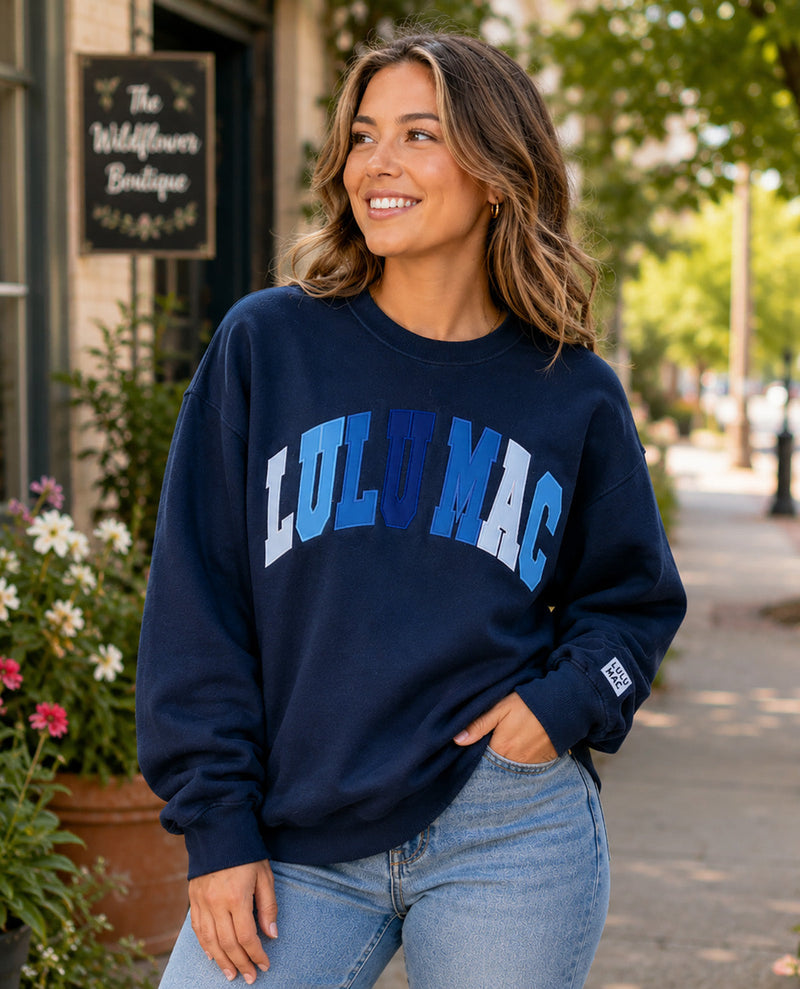 A woman wearing a navy blue crewneck sweatshirt with 'LULU MAC' printed in blue and light blue block letters across the chest, paired with light wash blue jeans, standing outdoors on a sidewalk.