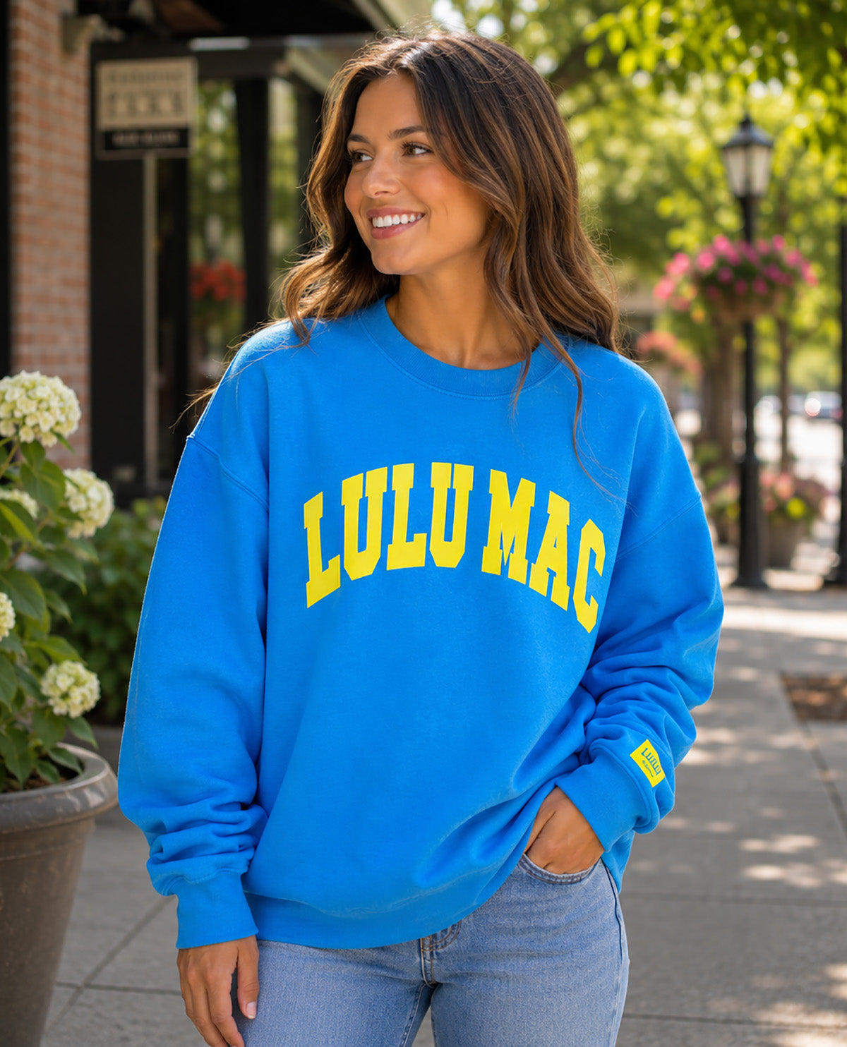A woman wearing a bright blue crewneck sweatshirt with 'LULU MAC' printed in yellow arched letters across the chest, paired with light blue jeans, standing on a sunny sidewalk.