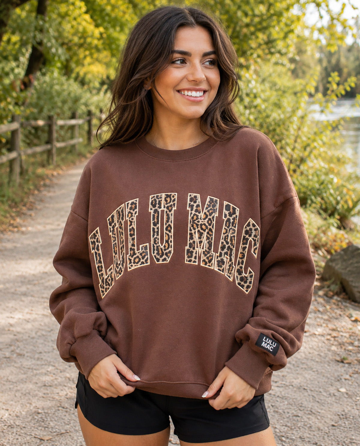 A woman wearing a brown crewneck sweatshirt with 'LULUMIC' printed in leopard-print letters, standing on an outdoor path with trees in the background.