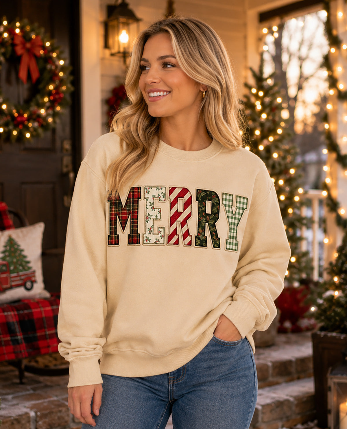 A woman wearing a cream-colored crewneck sweatshirt with 'MERRY' spelled out in festive patterned letters, paired with blue jeans, standing in a warmly lit holiday home setting.