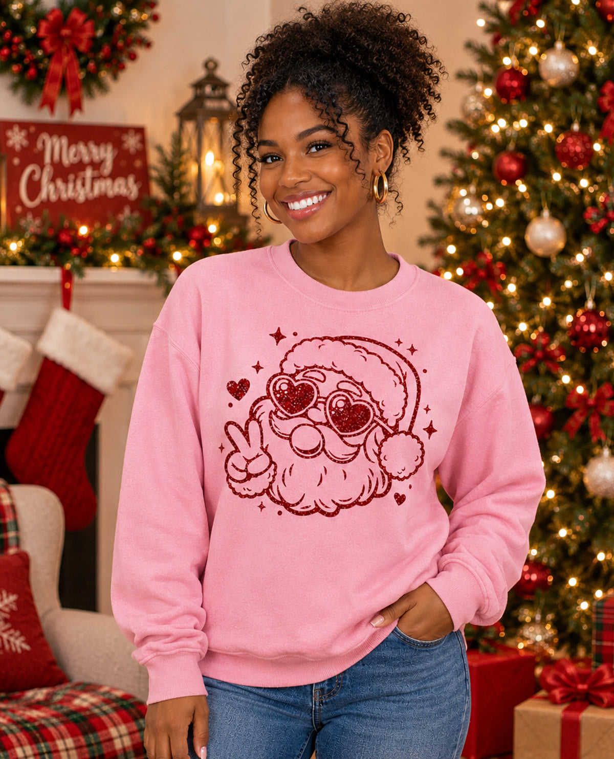 A woman wearing a pink crewneck sweatshirt with a red glittery Santa Claus graphic, posed in a cozy, decorated Christmas living room with a lit tree, stockings, and a fireplace.