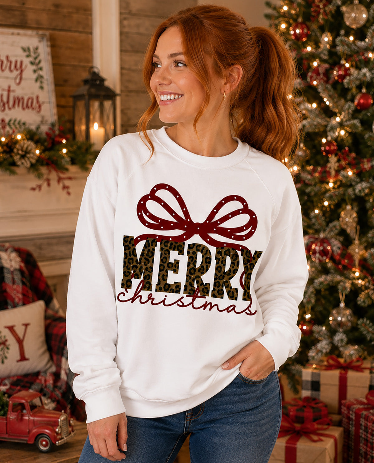 A woman wearing a white crewneck sweatshirt with a 'Merry Christmas' graphic featuring a red bow and leopard-print lettering, posed in a cozy holiday living room setting with a decorated Christmas tree in the background.