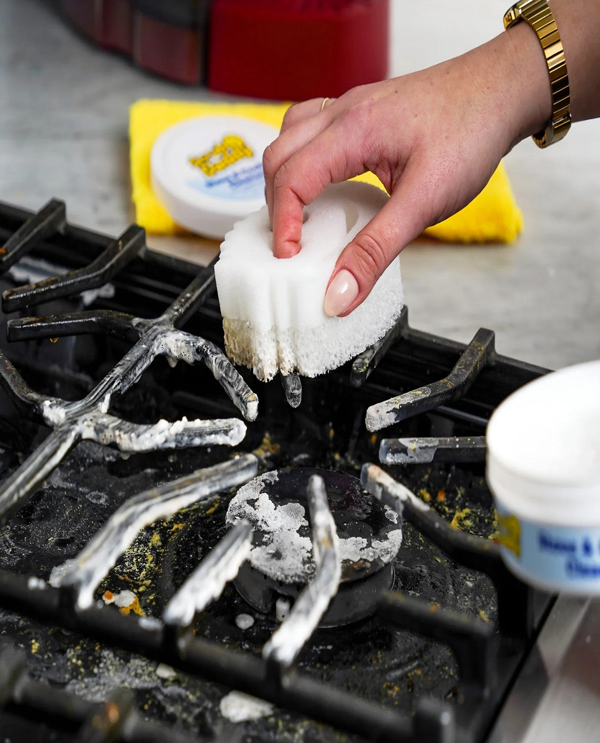 A hand wearing a gold bracelet holds a white cleaning sponge over a dirty black gas stove burner, with cleaning product containers and a yellow cloth in the background.