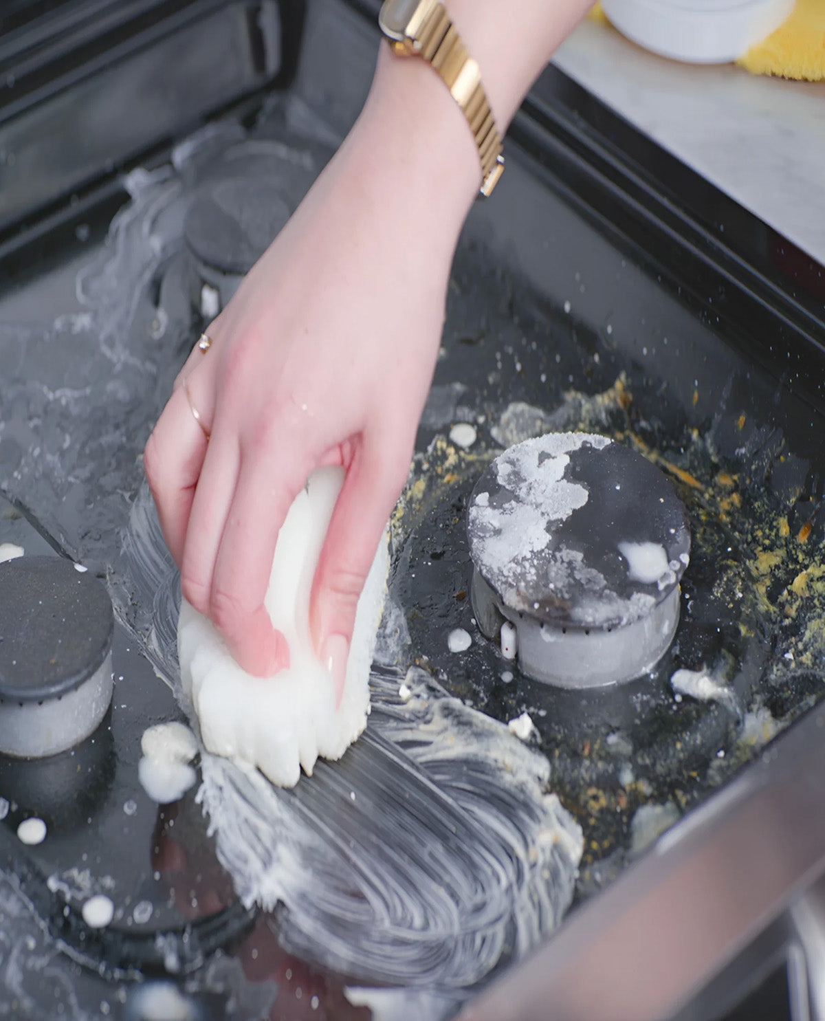 A close-up of a person's hand with a gold watch and ring scrubbing a dirty black stovetop burner with a white sponge, creating soapy foam.