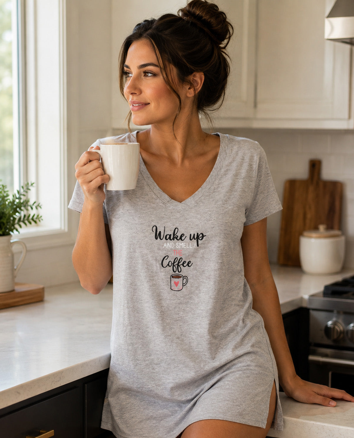 A woman in a gray heather V-neck t-shirt dress stands in a bright kitchen, holding a white mug. The dress features a graphic print that reads 'Wake up Coffee' with a small coffee cup and heart illustration.