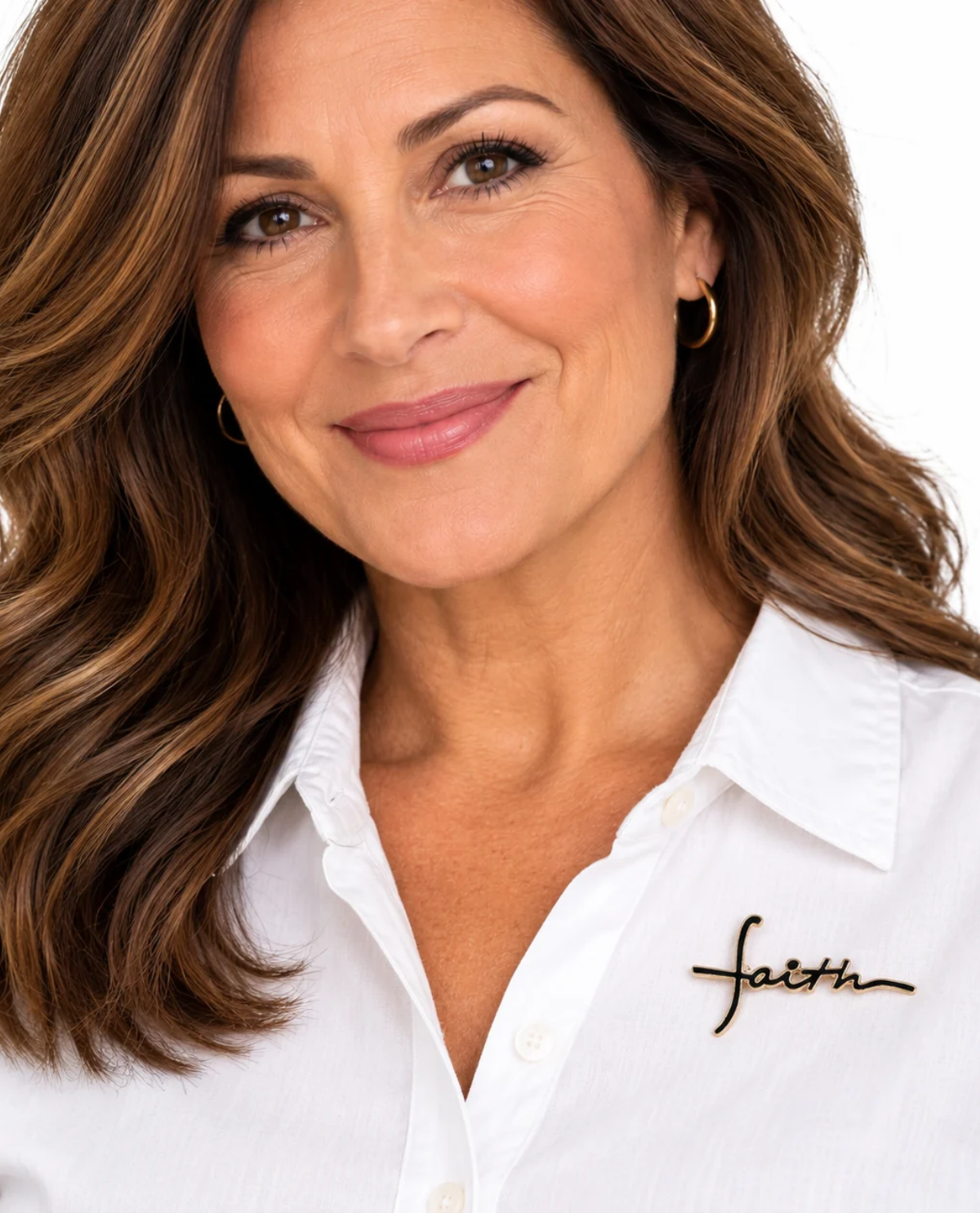 A close-up portrait of a smiling woman with brown hair wearing a white button-up shirt with a black script 'faith' pin on the chest, against a plain white background.