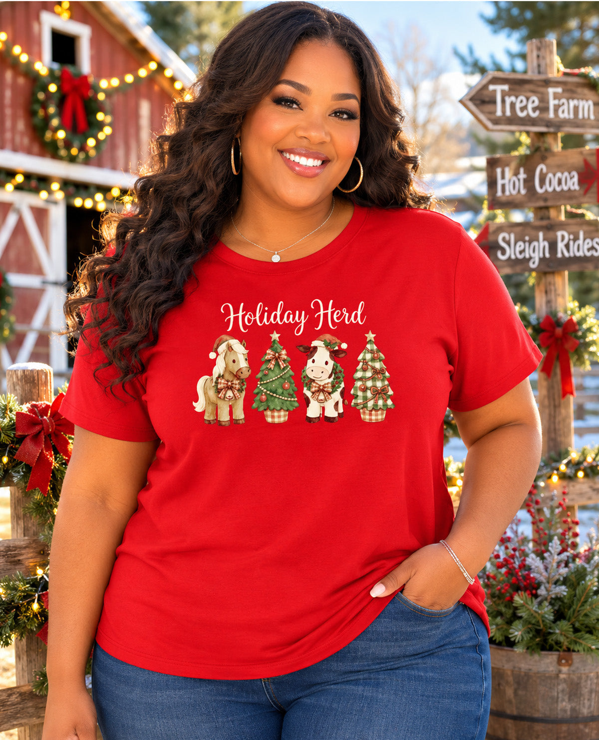A smiling woman wearing a bright red graphic t-shirt with 'Holiday Herd' and four holiday-themed animals printed on the front, posed in front of a rustic Christmas-themed barn background.