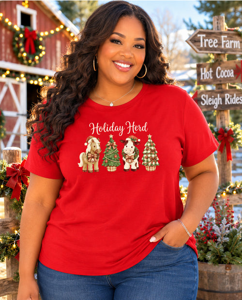A smiling woman wearing a bright red graphic t-shirt with 'Holiday Herd' and four holiday-themed animals printed on the front, posed in front of a rustic Christmas-themed barn background.