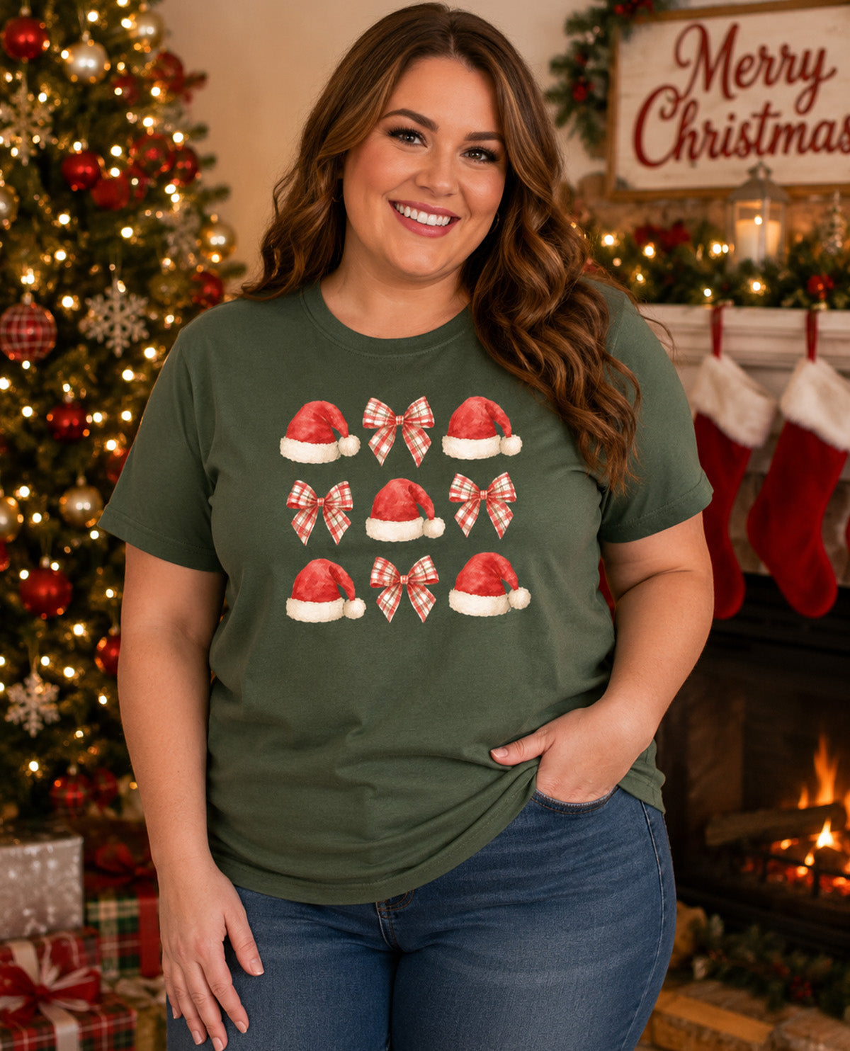 A woman wearing an olive green t-shirt with a graphic print of Santa hats and red-and-white bows, posed in a cozy holiday living room setting with a Christmas tree and fireplace in the background.