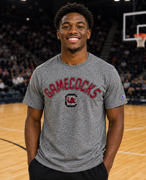 A smiling man standing on a basketball court wearing a heather gray Champion t-shirt with 'GAMECOCKS' and a red logo printed on the front.