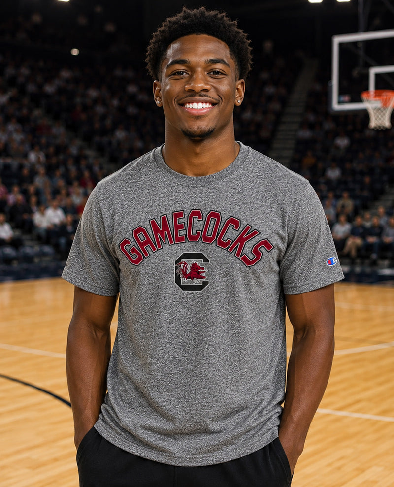 A smiling man standing on a basketball court wearing a heather gray Champion t-shirt with 'GAMECOCKS' and a red logo printed on the front.