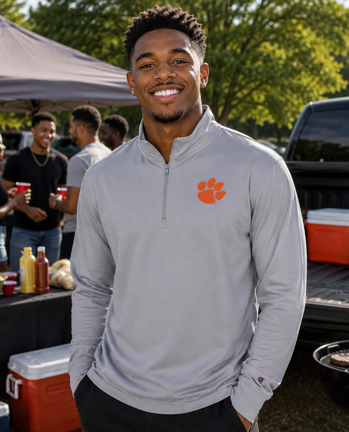 A smiling man wearing a light gray quarter-zip pullover with an orange Clemson Tigers paw logo on the left chest, standing outdoors at a tailgate or barbecue with people and coolers in the background.