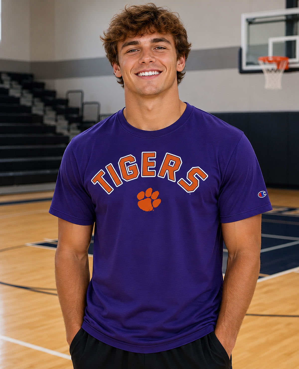 A smiling young man stands on a basketball court wearing a purple short-sleeve T-shirt with 'TIGERS' and an orange paw print on the front, plus a small Champion logo on the sleeve.