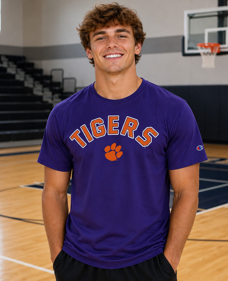 A smiling young man stands on a basketball court wearing a purple short-sleeve T-shirt with 'TIGERS' and an orange paw print on the front, plus a small Champion logo on the sleeve.