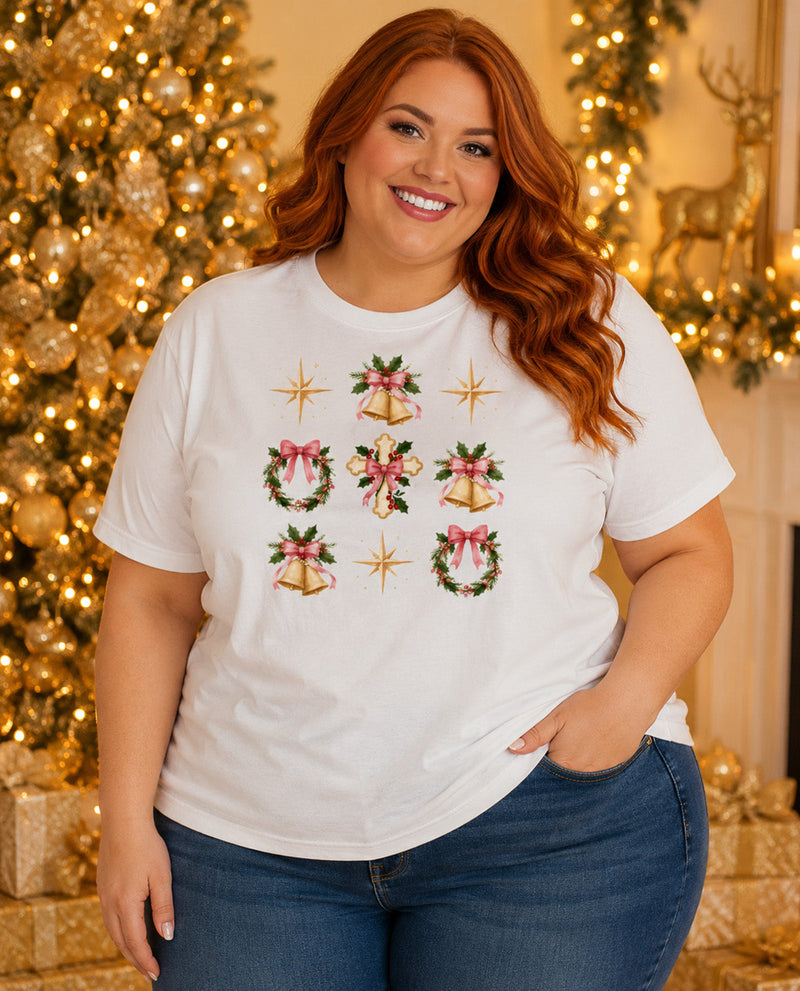 A woman wearing a white short-sleeve t-shirt with a Christmas-themed graphic print featuring bells, wreaths, bows, and stars, posed in front of a warmly lit, decorated Christmas tree.