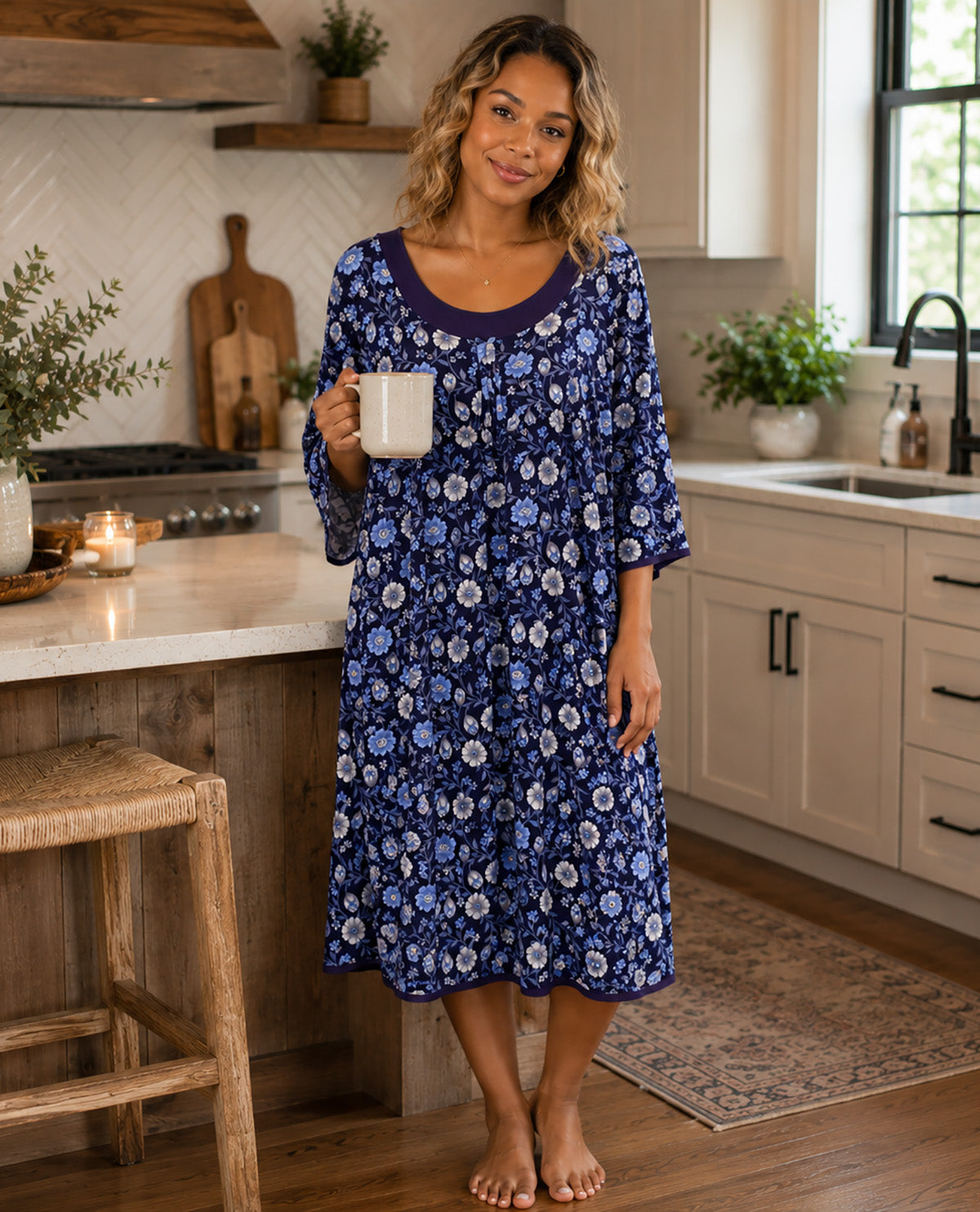 A woman stands barefoot in a bright kitchen wearing a blue floral nightgown with three-quarter sleeves and holding a white mug.
