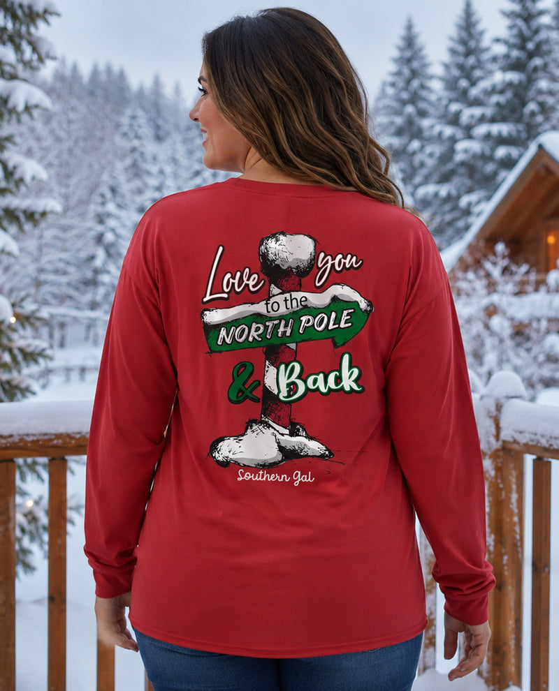 A woman wearing a red long-sleeve shirt with a large back graphic that reads 'Love you to the NORTH POLE & Back' with a snowman and boots, photographed outdoors in a snowy forest setting.