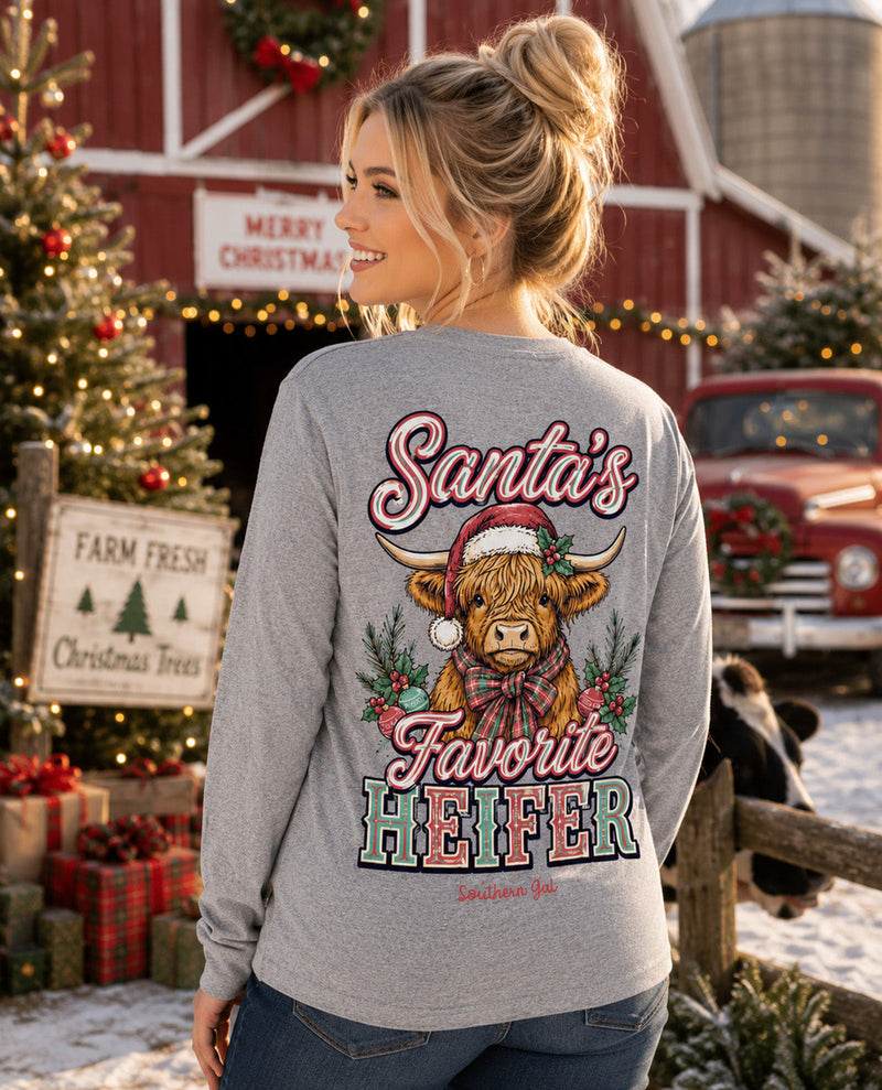 A woman stands outdoors in a snowy farm setting wearing a heather gray long-sleeve shirt with a large Christmas graphic on the back. The graphic shows a cartoon Highland cow in a Santa hat with the text 'Santa's Favorite Heifer.