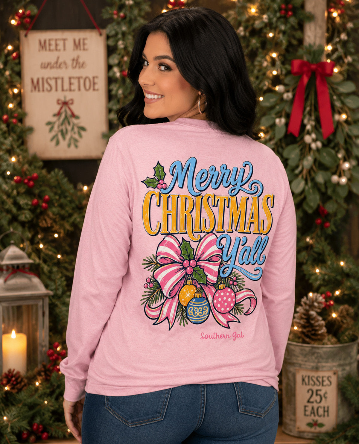 A woman wearing a light pink long-sleeve shirt with a large 'Merry Christmas Y'all' graphic on the back, posed in front of a decorated Christmas tree.