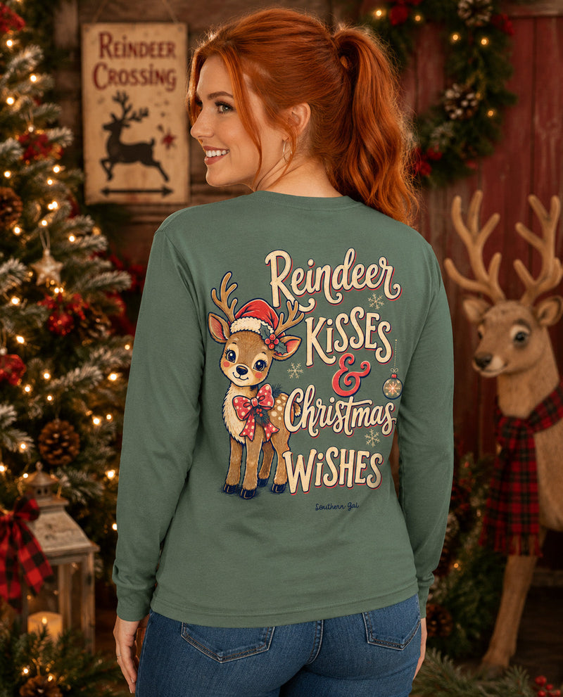 A woman with red hair is shown from the back wearing a sage green long-sleeve shirt with a festive Christmas graphic. The graphic features a cartoon deer wearing a Santa hat and scarf with the text 'Reindeer Kisses & Christmas Wishes' in cream and gold lettering.