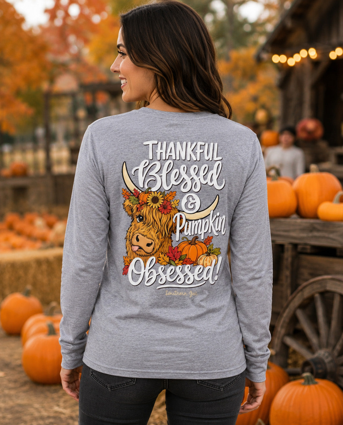 A woman shown from the back wearing a heather gray long-sleeve t-shirt with a large autumn graphic on the back. The graphic features a cartoon Highland cow surrounded by pumpkins and fall leaves, with the text 'THANKFUL Blessed & Pumpkin Obsessed!'.