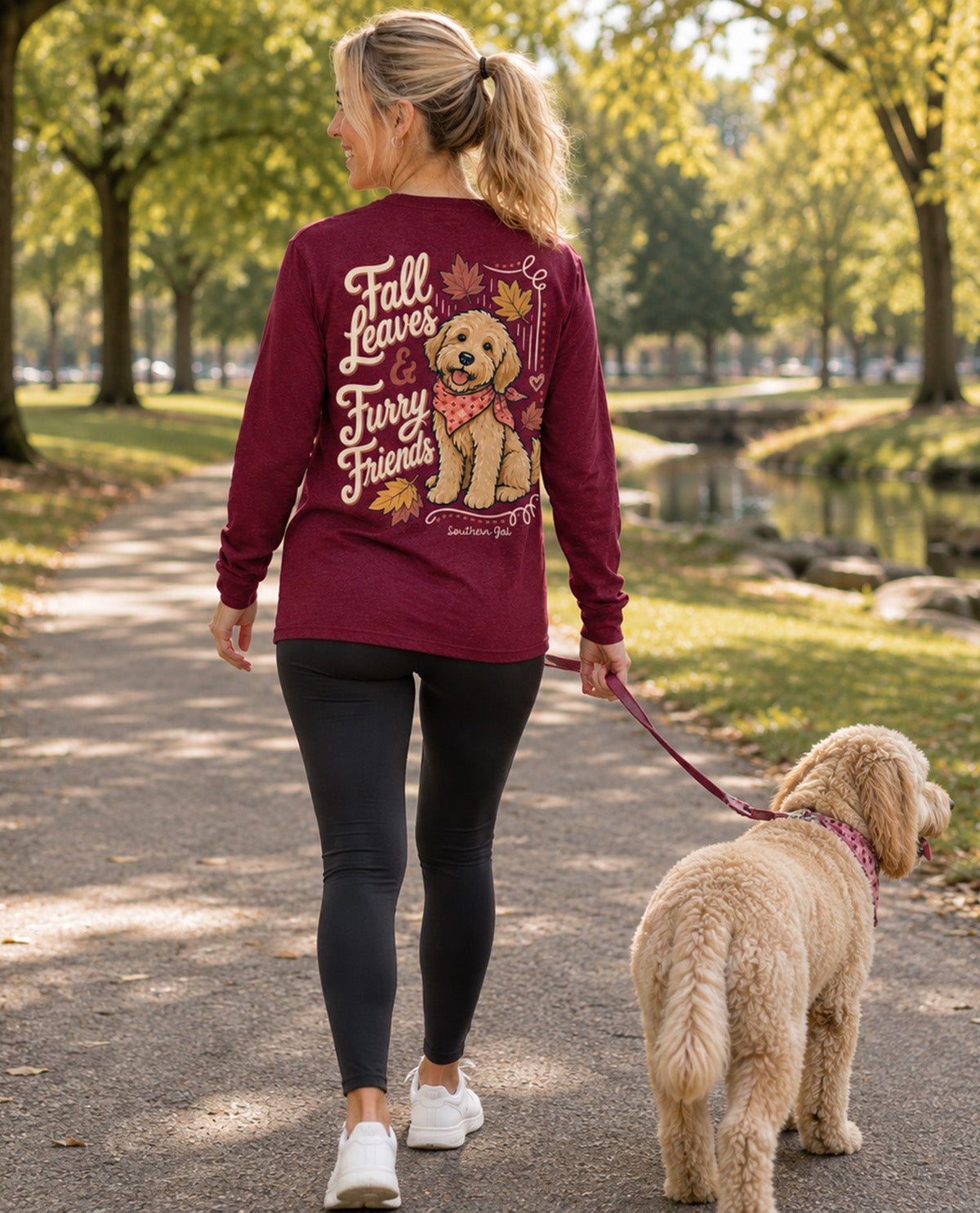 A woman walking a goldendoodle dog on a leash along a park path, wearing a burgundy long-sleeve shirt with a large graphic of a golden retriever and the text 'Fall Leaves furry friends' on the back.