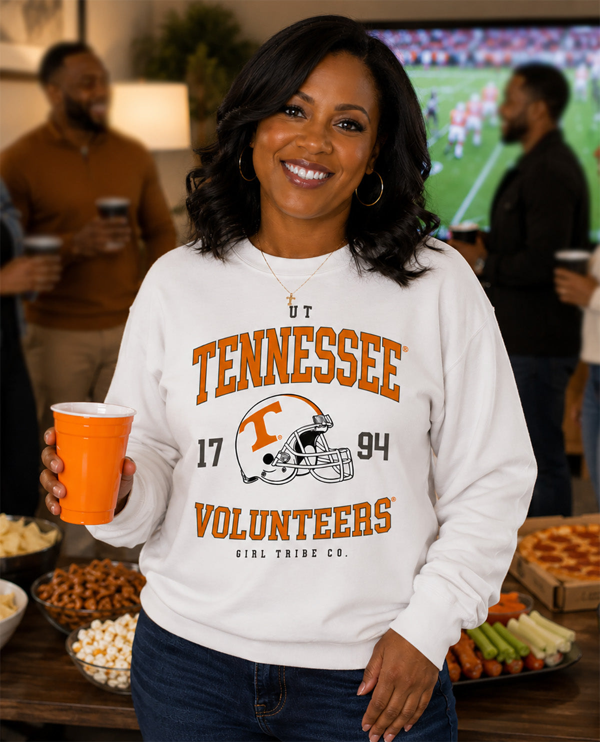 A smiling woman stands in a social setting wearing a white long-sleeve sweatshirt with a large Tennessee Volunteers graphic on the front. The graphic features orange lettering, the number 17, the year 94, and a football helmet logo.