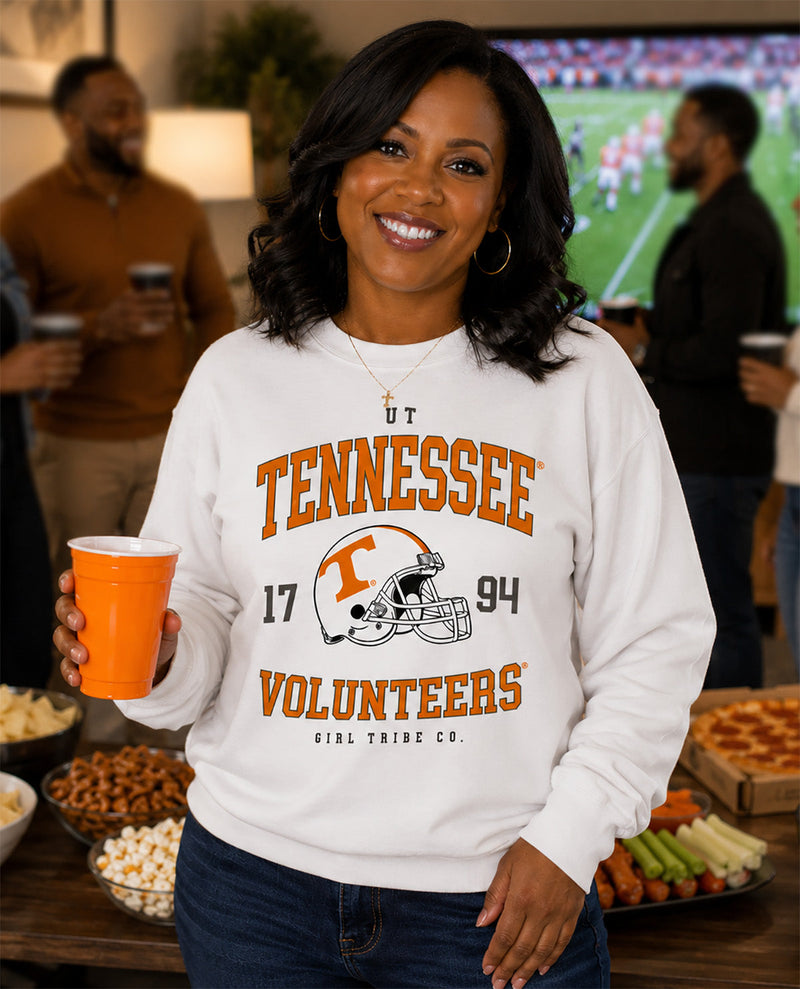 A smiling woman stands in a social setting wearing a white long-sleeve sweatshirt with a large Tennessee Volunteers graphic on the front. The graphic features orange lettering, the number 17, the year 94, and a football helmet logo.