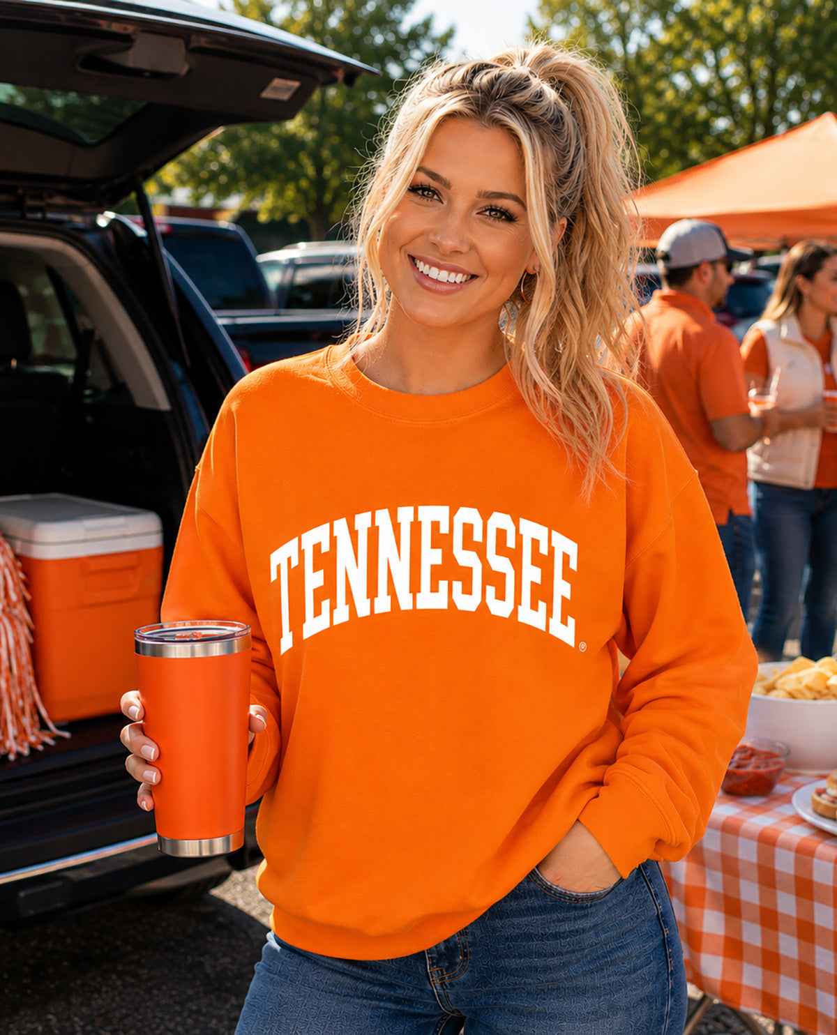 A woman wearing an orange crewneck sweatshirt with 'TENNESSEE' printed in white arched letters, holding an orange tumbler at an outdoor tailgate or market event.