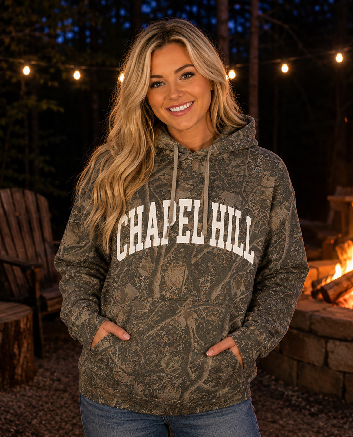 A woman wearing a camouflage hoodie with 'CHAPEL HILL' printed in white arched letters across the chest, standing outdoors at night near a fire pit and string lights.