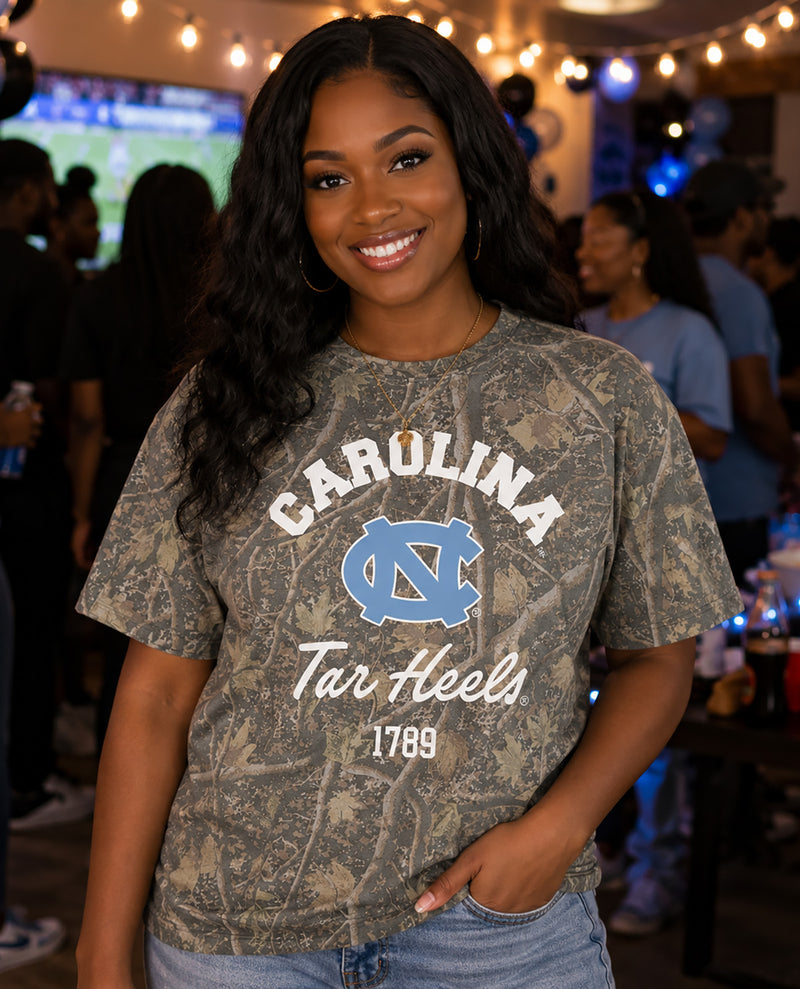 A woman smiling at a social event wearing a faded camouflage Carolina Tar Heels graphic t-shirt with a blue and white UNC logo.