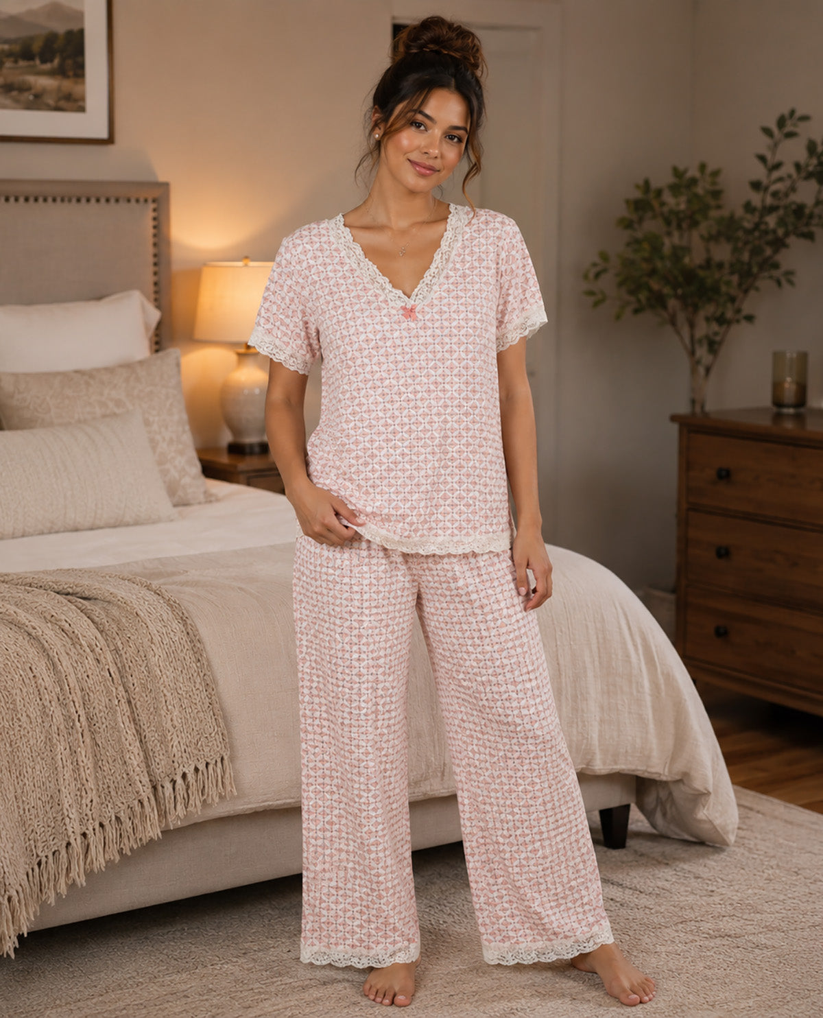 A woman stands barefoot in a bedroom wearing a light pink and white patterned two-piece pajama set with a V-neck top and wide-leg pants.