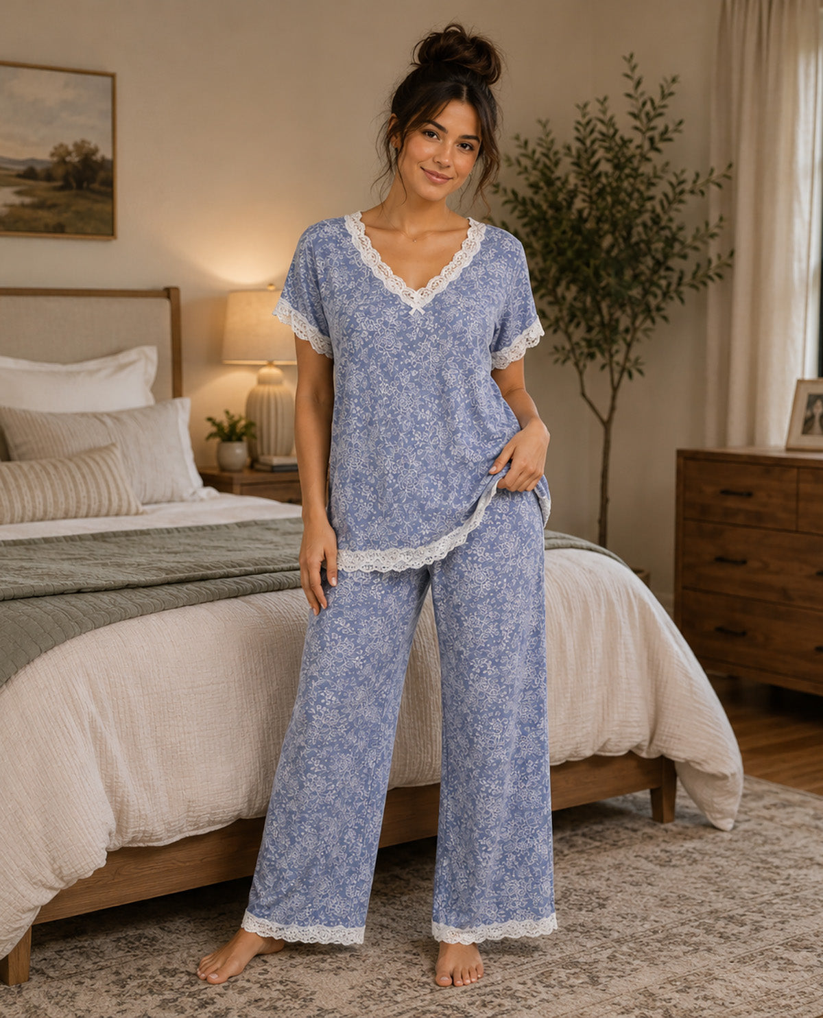 A woman stands barefoot in a bedroom wearing a light blue floral pajama set with white lace trim. The set includes a short-sleeve V-neck top and matching wide-leg pants.