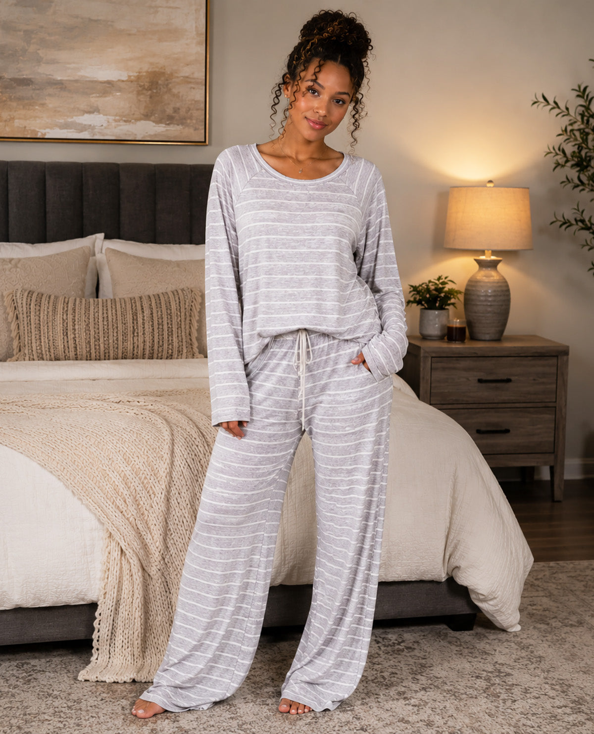 A woman stands barefoot in a bedroom wearing a light gray and white striped lounge set consisting of a long-sleeve top and matching wide-leg pants.