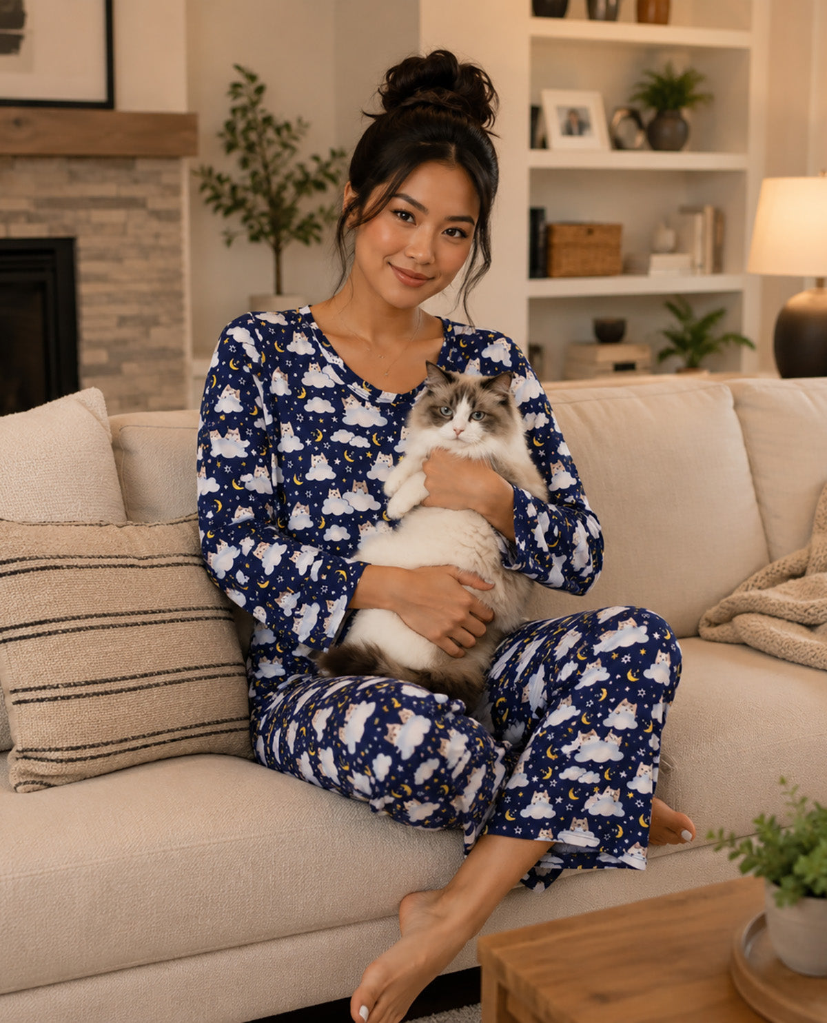 A woman sitting on a beige couch in a cozy living room, holding a white and gray cat. She is wearing a matching navy blue and white pajama set with a small yellow star pattern.