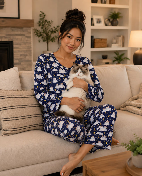 A woman sitting on a beige couch in a cozy living room, holding a white and gray cat. She is wearing a matching navy blue and white pajama set with a small yellow star pattern.