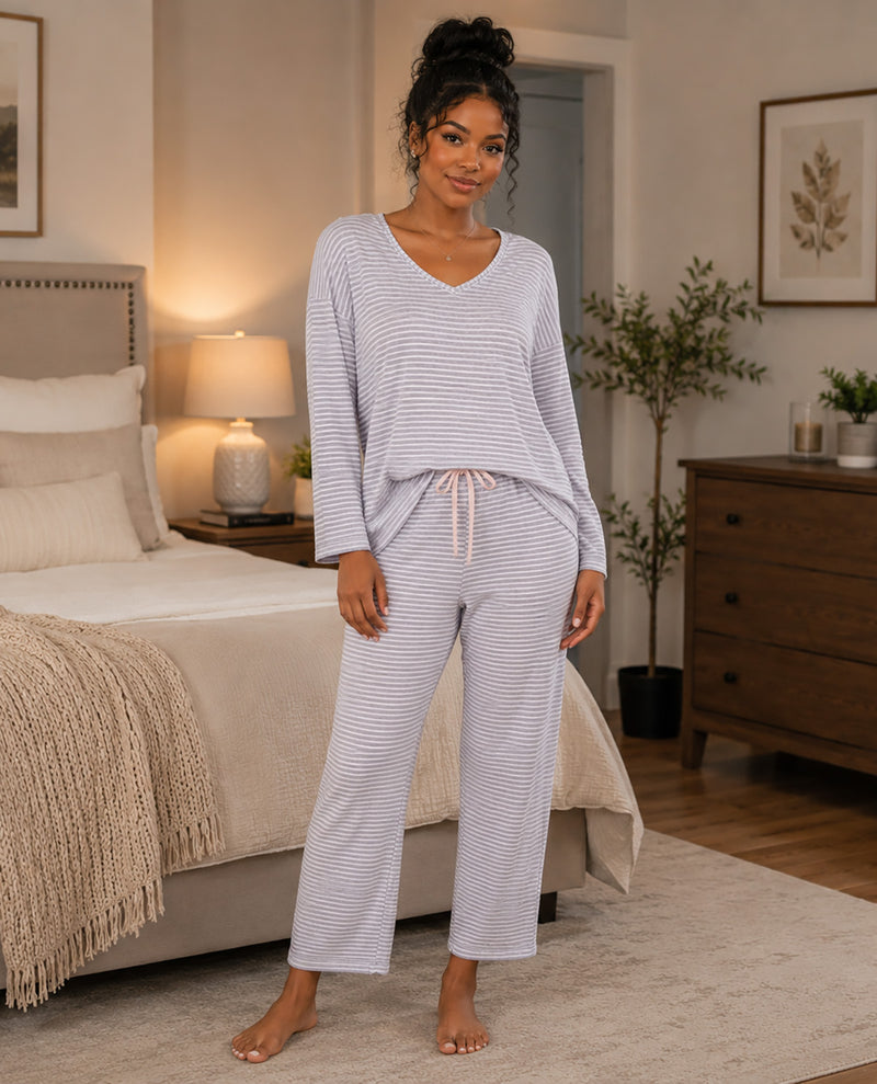 A woman stands barefoot in a bedroom wearing a light lavender and white striped pajama set with a V-neck long-sleeve top and matching drawstring pants.