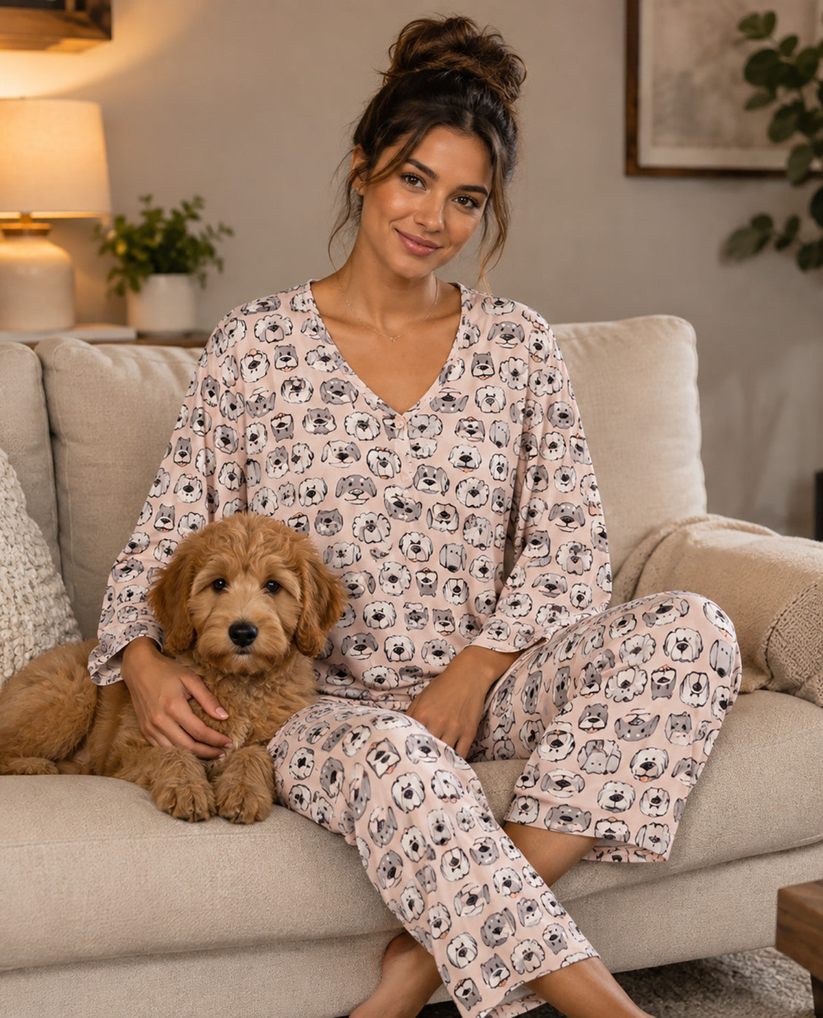 A woman wearing a matching long-sleeve pajama set with a dog face print sits on a beige couch holding a small golden dog. The pajamas are light pink with a repeating black and gray dog face pattern.