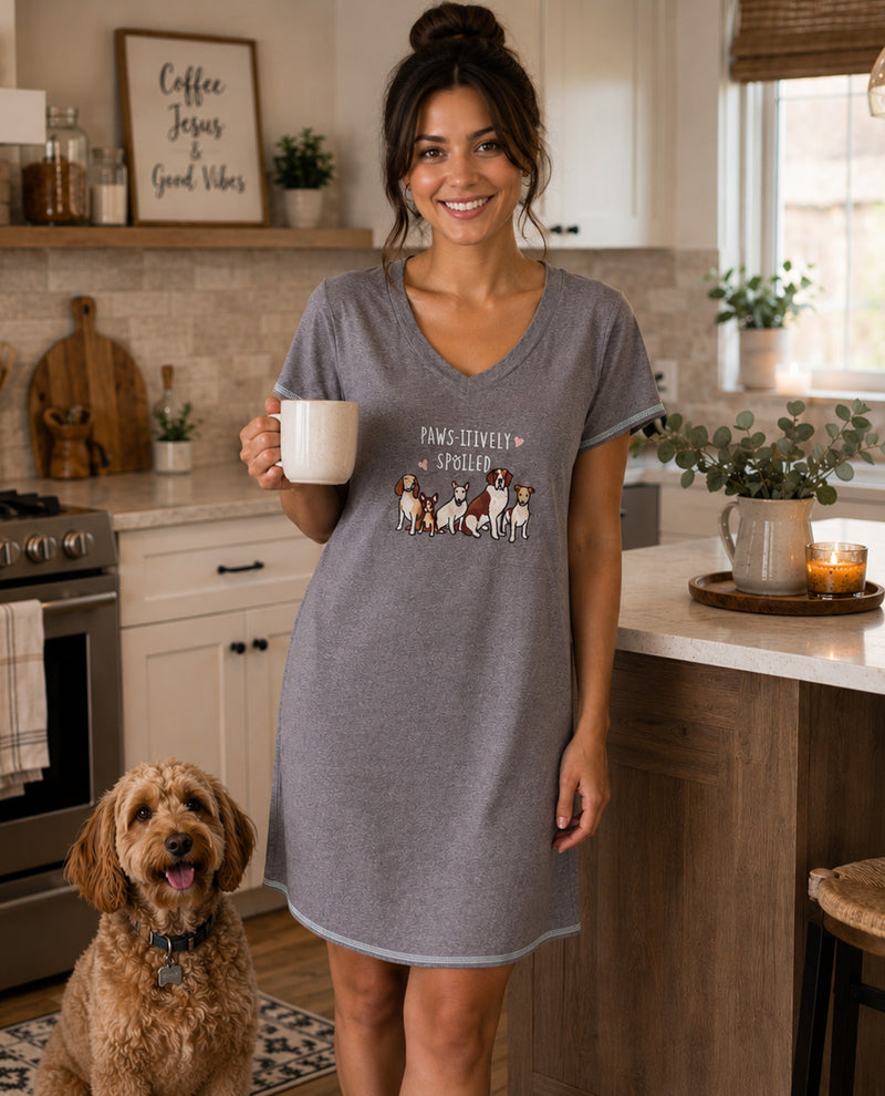 A woman wearing a heather gray V-neck sleep shirt dress stands in a kitchen holding a white mug, with a small graphic of dogs and the text 'Pawfectly Happy Dogs' on the front. A brown poodle dog sits on the floor nearby.