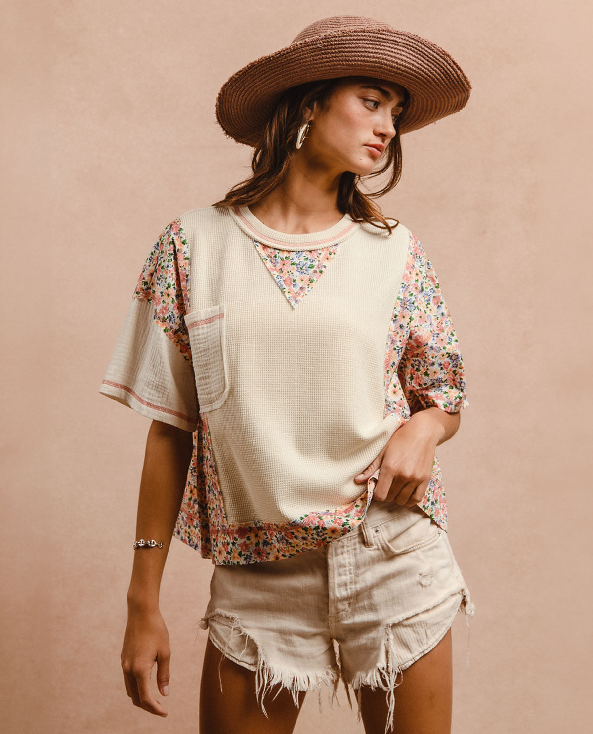 A woman wearing a cream-colored short-sleeve top with colorful floral patterned side panels and a matching floral triangle neckline, paired with distressed denim shorts and a wide-brimmed straw hat, set against a solid beige background.