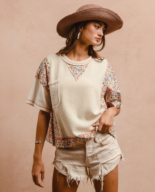 A woman wearing a cream-colored short-sleeve top with colorful floral patterned side panels and a matching floral triangle neckline, paired with distressed denim shorts and a wide-brimmed straw hat, set against a solid beige background.