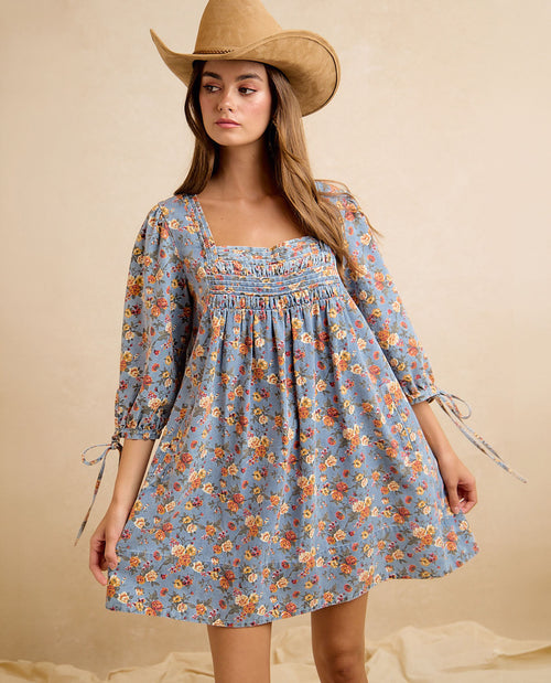 A woman wearing a light blue floral mini dress with a smocked bodice, square neckline, and tie details at the cuffs, styled with a tan cowboy hat against a warm beige studio backdrop.