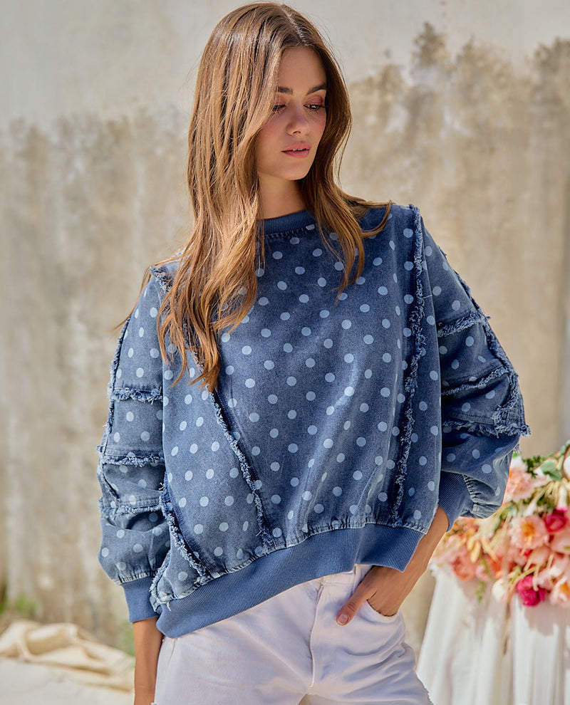 A woman wearing a blue denim-style sweatshirt with white polka dots and distressed, frayed details on the sleeves, paired with white pants, set against a softly blurred rustic background.