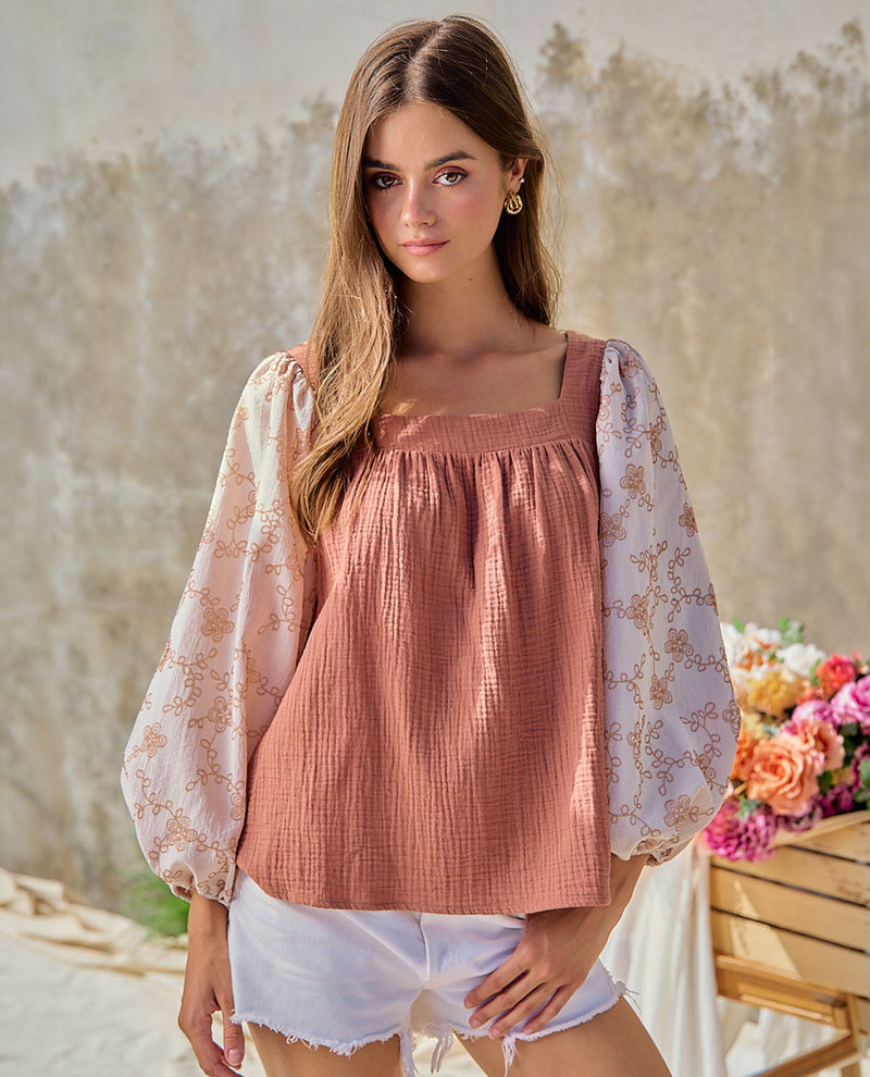 A woman wearing a dusty rose puff-sleeve top with a white floral overlay and white shorts, standing in front of a textured beige wall with a flower basket in the background.