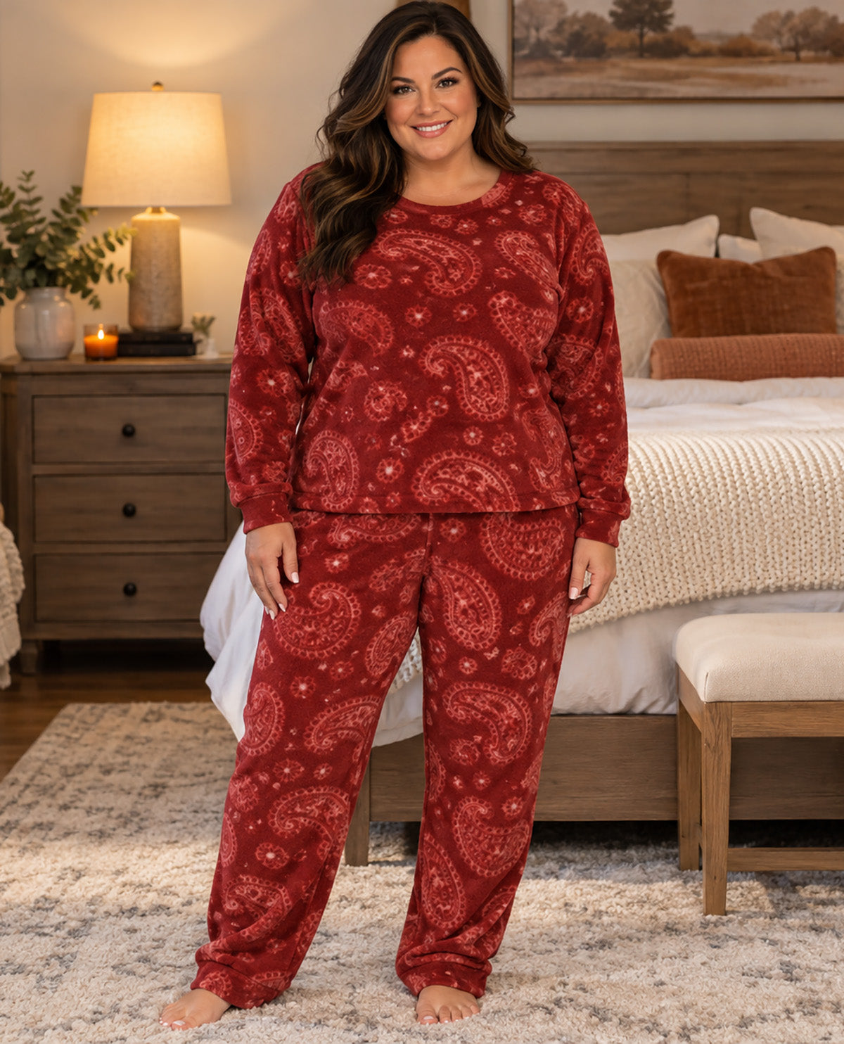 A woman stands barefoot in a bedroom wearing a matching long-sleeve pajama top and pants set in a deep red paisley pattern. The bedroom background features a bed, bedside table with a lit lamp, and a neutral-toned rug.