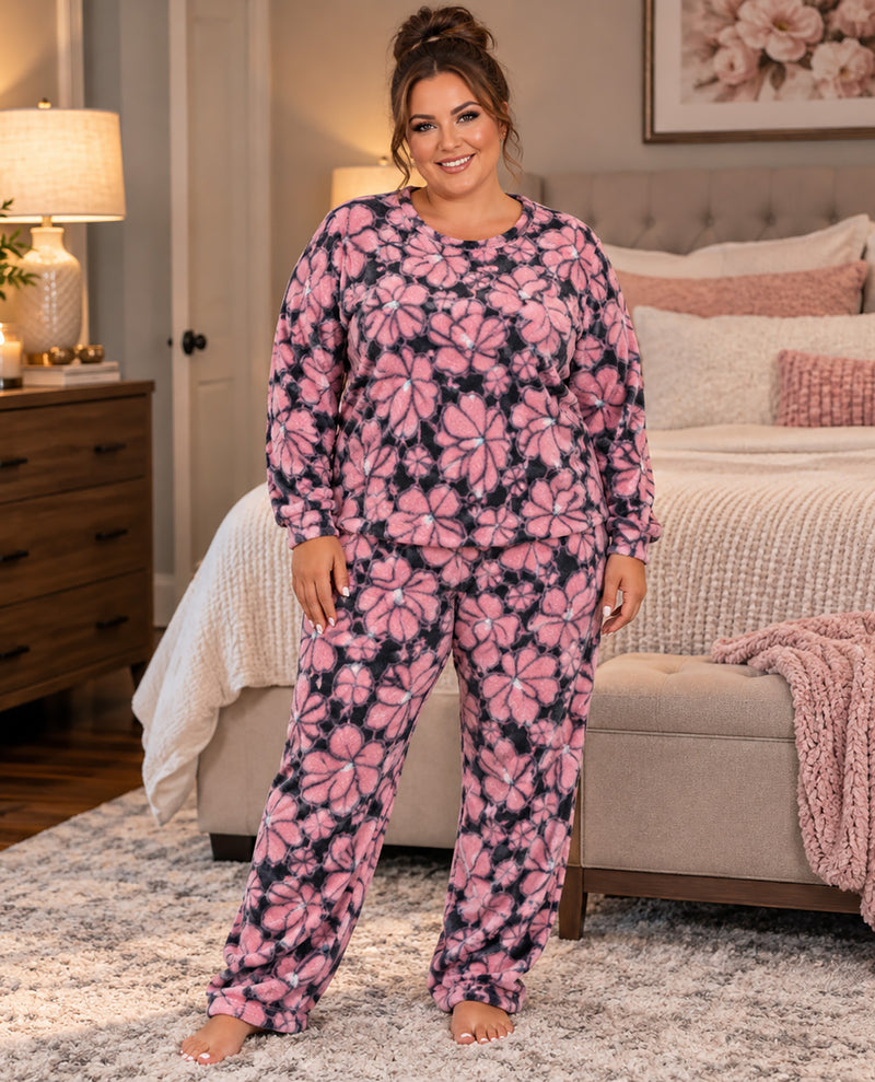 A woman stands in a bedroom wearing a matching long-sleeve pajama set with a pink and black floral pattern. The set includes a crewneck top and matching pants.