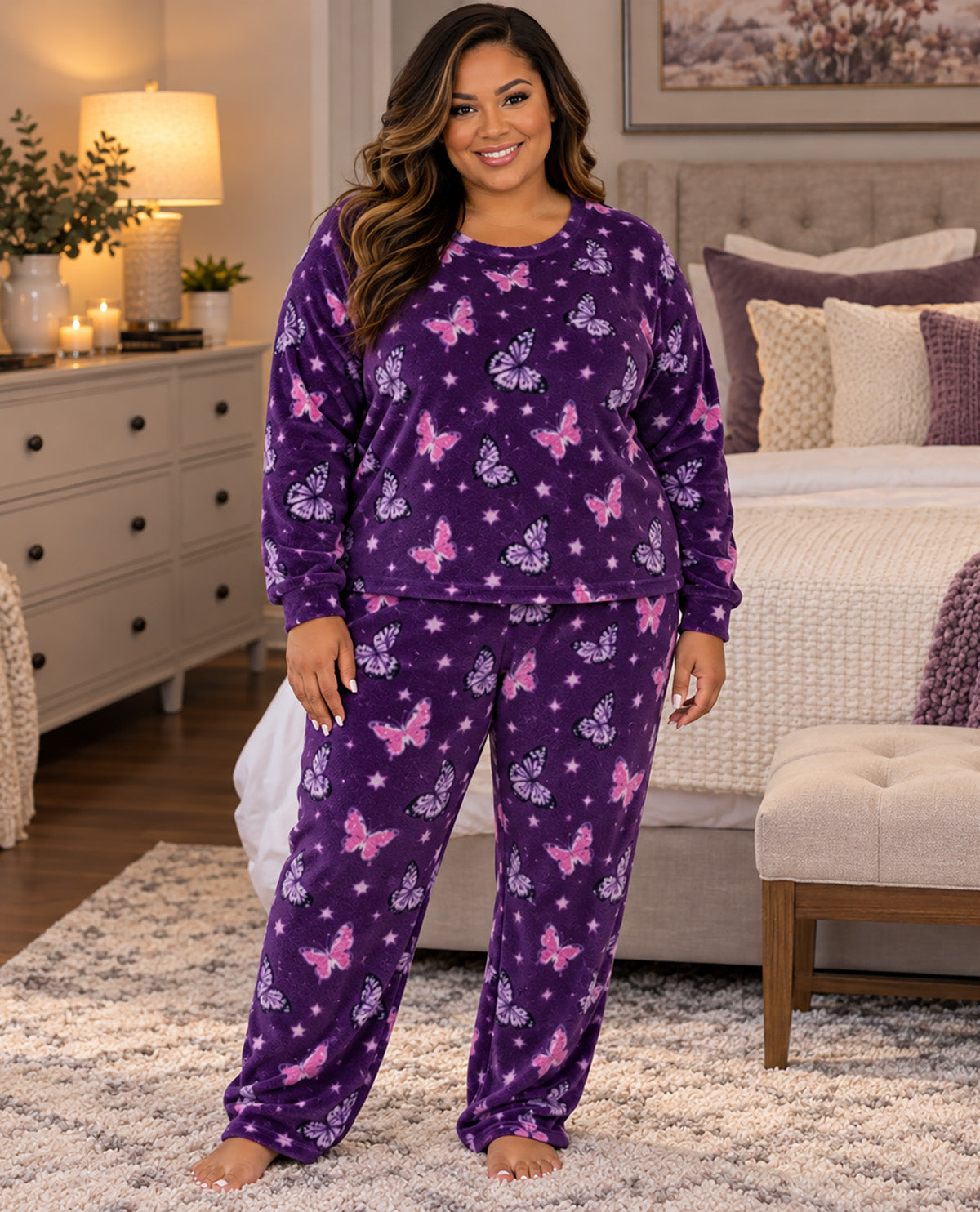 A woman stands barefoot in a bedroom wearing a matching purple pajama set with a pink and lavender butterfly and star print. The set includes a long-sleeve top and matching pants.